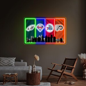 May include: A rectangular neon wall sign featuring four vertical color panels in green, blue, red, and orange. The sign displays the Philadelphia Eagles, Phillies, 76ers, and Flyers logos above a black silhouette of the Philadelphia city skyline.
