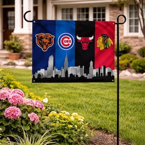 May include: A garden flag displaying the logos of the Chicago Bears, Chicago Cubs, Chicago Bulls, and Chicago Blackhawks. The flag has a black skyline at the bottom. The flag is displayed in a garden with pink flowers.