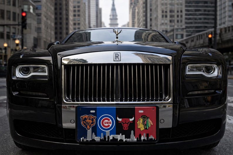 May include: A black Rolls-Royce with a silver grille and hood ornament. The number plate features logos for the Chicago Bears, Cubs, Bulls, and Blackhawks, with a cityscape silhouette. The car is parked on a city street.