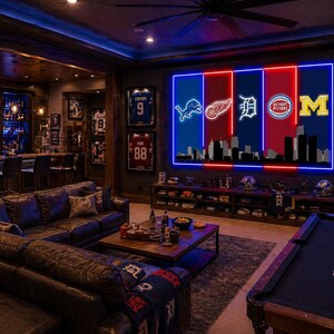 May include: A large rectangular wall art piece featuring five vertical illuminated panels with neon-style logos for the Detroit Lions, Red Wings, Tigers, Pistons, and the University of Michigan, set above a dark silhouette of the Detroit city skyline.
