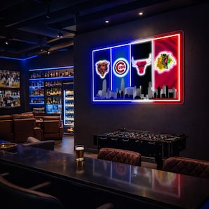 May include: A rectangular wall-mounted neon sign featuring the Chicago Bears, Cubs, Bulls, and Blackhawks logos arranged horizontally above a silhouette of the Chicago skyline, illuminated in vibrant blue, white, black, and red neon light tubes.