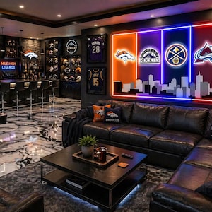 May include: A large rectangular wall-mounted neon sign featuring the logos of the Denver Broncos, Colorado Rockies, Denver Nuggets, and Colorado Avalanche above a white city skyline silhouette, set against a dark wall in a finished basement.