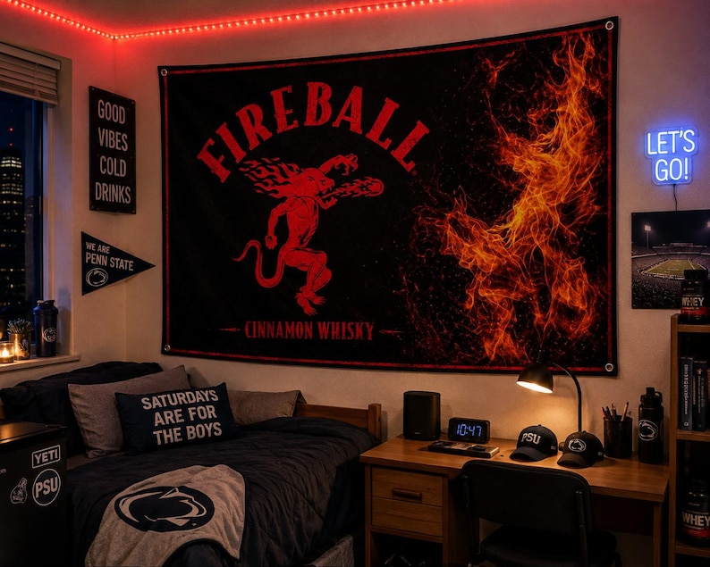 K&ouml;nnte beinhalten: Ein schwarzes Fireball Cinnamon Whisky Banner mit roter Teufelsgrafik und Flammen. Der Raum umfasst einen Schreibtisch mit Digitaluhr, Penn State Kappen und einem Neon-Schild "Let's Go!". Der Raum ist mit roten Lichtern beleuchtet.