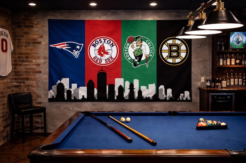 Pode incluir: Uma grande bandeira de parede com os logotipos das equipas desportivas de Boston: Patriots, Red Sox, Celtics e Bruins. A bandeira &eacute; dividida em quatro sec&ccedil;&otilde;es com um horizonte a preto e branco na parte inferior. Uma mesa de bilhar com feltro azul e tacos est&aacute; em primeiro plano.