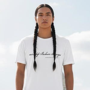 Puede incluir: Una persona con una camiseta blanca con el texto "my life Indian changes" en escritura negra, combinada con vaqueros azul oscuro y zapatillas blancas. El fondo presenta un gran edificio blanco con una cúpula.