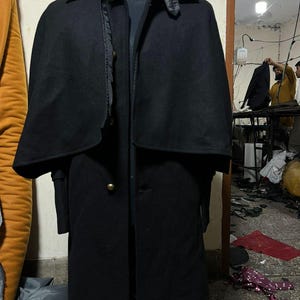 May include: A black wool coat with a cape-like shoulder design, displayed on a mannequin. The coat features a button-down front and a classic collar. The garment is a dark, solid color, and appears to be a winter coat.