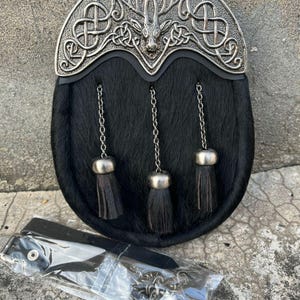 Puede incluir: Un sporran negro con una parte superior de metal plateado con un diseño de cabeza de ciervo y nudo celta. El sporran tiene tres borlas y una cadena. Se incluyen accesorios adicionales.