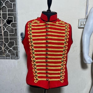 Può includere: Un gilet rosso in stile militare con finiture e bottoni dorati. Il gilet ha un colletto alto ed è esposto su un manichino nero. Le finiture dorate sono disposte in file orizzontali sul petto e sulle spalle.