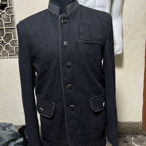 Könnte beinhalten: Dunkelgraue Wolljacke mit Stehkragen und dekorativen Knöpfen. Die Jacke hat zwei Vordertaschen und einen grauen Kragen. Die Jacke wird auf einer Schaufensterpuppe präsentiert.
