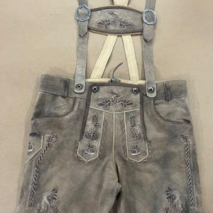 Könnte beinhalten: Graue Lederhose-Shorts mit Hosenträgern. Die Shorts haben dekorative Stickereien auf den Taschen und der Frontplatte. Die Hosenträger sind beige mit silbernen Schnallen und einem dekorativen Rückenteil aus Leder.