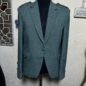Puede incluir: Un blazer de tweed gris con cierre de un solo botón, charreteras y botones decorativos en las mangas. La chaqueta se exhibe en un maniquí. El blazer tiene dos bolsillos delanteros y solapas con muescas.