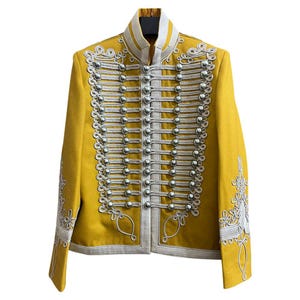 Puede incluir: Chaqueta de estilo militar amarillo mostaza con botones plateados y ribetes decorativos plateados. La chaqueta presenta un cuello alto y bordados ornamentados en las mangas. Prenda de abrigo única.