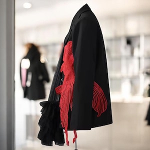 Puede incluir: Un blazer negro con un llamativo diseño rojo en forma de pez en el lateral. El blazer presenta un detalle único de tela negra en capas en la parte delantera y se exhibe sobre un soporte metálico. El diseño es llamativo y artístico.
