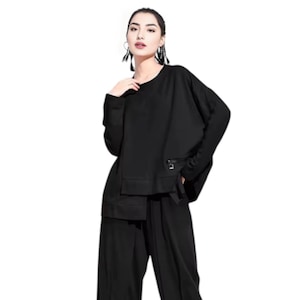 Puede incluir: Un conjunto negro de dos piezas con una parte superior de manga larga y pantalones de pernera ancha. La parte superior tiene cuello redondo, un pequeño bolsillo con cremallera y un corte holgado. Los pantalones son de cintura alta y fluidos.