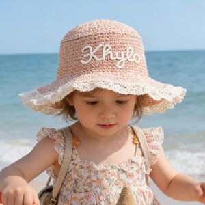 May include: A child wearing a pink sun hat with the name "Khylo" embroidered on it, playing on a sandy beach. The hat has a white lace trim. The child is wearing a floral dress and using a yellow shovel to build a sandcastle.