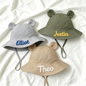 Könnte beinhalten: Drei Kinder-Eimerhüte in Grau, Olivgrün und Beige. Jeder Hut hat ein Paar kleiner, abgerundeter Ohren oben und einen Kinnriemen. Die Namen "Elliot", "Justin" und "Theo" sind auf die Vorderseite jedes Hutes gestickt.