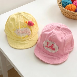 Op de afbeelding: Twee baseballpetten voor kinderen. Eén is geel met de naam "Holly" geborduurd op een patch en een roze snoepvormige versiering. De andere is roze met de naam "Ida" geborduurd op een patch.