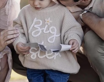 May include: A beige long-sleeve knit sweater featuring the white embroidered text "Big Sister" across the chest, accented with a small white flower detail. The sweater has a ribbed texture and is paired with dark blue denim jeans.