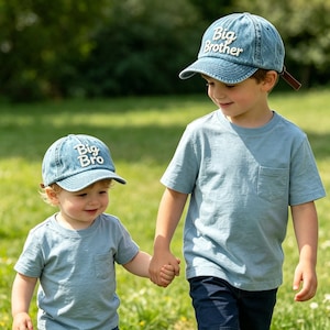 Op de afbeelding: Twee kinderen dragen bijpassende blauwe denim baseballpetten en lichtblauwe t-shirts. Op de ene pet staat "Big Brother" en op de andere "Big Bro" in witte letters. De kinderen houden elkaars hand vast en lopen op gras.