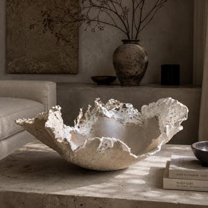 Pode incluir: Uma taça grande e irregular com um exterior texturizado em branco sujo e um interior liso de cor clara. O rebordo apresenta uma forma orgânica irregular com arestas porosas semelhantes a crostas que contrastam com a base profunda.