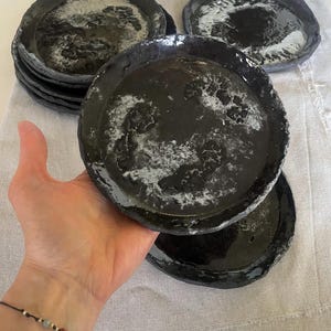 May include: A set of circular ceramic plates with a dark black finish and a glossy, textured surface featuring white marbled accents. Each plate has an uneven, hand-formed rim and a shallow basin, showcasing a distinct, organic, and rustic appearance.