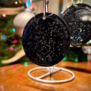 May include: Black circular ornament with the text "Ovation - Chicago IL" and "January 18, 2026". The ornament has a constellation design and the names "Jill & Jonathan". The ornament is hanging on a white metal stand.