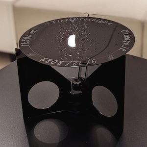 May include: A black metal prototype stand with a round top that says "First Prototype" and "Chicago, IL". The top also has the date "6/12/2023" and the time "11:59 p.m.".