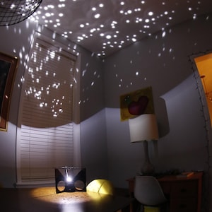 May include: A black projector casts a starry night sky pattern on the ceiling of a room. The projector is on a table with a yellow cushion and a white chair. A white lamp with a white shade is in the background.