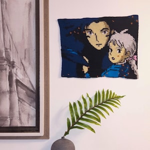 Könnte beinhalten: Ein rechteckiger Wandbehang aus Häkelarbeit mit einer verpixelten Grafik von zwei animierten Figuren. Eine Figur hat dunkles Haar und einen dunkelblauen Umhang, die andere helles Haar und ein blaues Hemd. Das Stück verwendet dunkelblaues, schwarzes, beiges und weißes Garn.