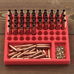 May include: A red reloading block with multiple holes holding brass casings. The block also has compartments for bullets and primers. The brass casings and bullets are a copper color, and the primers are gold.