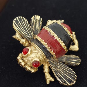 Puede incluir: Un broche de abeja de tono dorado con alas y cuerpo de esmalte negro y rojo. La abeja tiene ojos de pedrería roja.