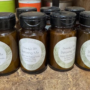 May include: Six amber glass jars with black lids filled with a detoxing mix. Each jar has a white label with green text that reads "Dowdy's DIY Detoxing Mix" and lists the ingredients: Epsom Salt, Baking Soda, Bentonite Clay, and Zendocrine Oil.