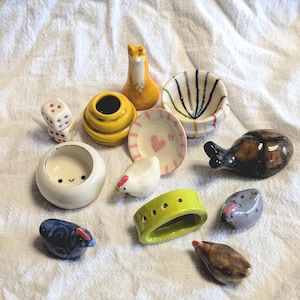 May include: A collection of small, hand-painted ceramic figurines. The assortment includes a fox, dice, bowls, a hen, and other whimsical shapes in various colors like yellow, white, blue, and brown. The objects are arranged on a white cloth.