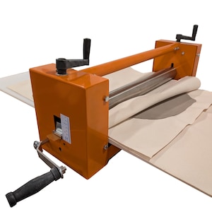 May include: An orange clay roller machine with a black handle. The machine has a long roller and a crank handle. Beige fabric and a grey clay piece are visible on the machine.