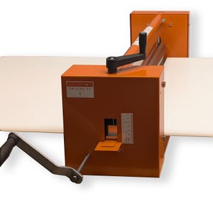 May include: An orange paper cutter with a black handle and a ruler on the side. The machine has a crank and a lever. The text "DOLCE CERAMICS DRAGON 21" is visible on the front.