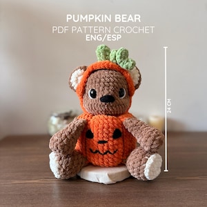 Puede incluir: Un oso de ganchillo marrón con un disfraz de calabaza naranja y un tallo verde en la cabeza. El oso tiene orejas y almohadillas blancas. El texto dice PUMPKIN BEAR PDF PATTERN CROCHET ENG/ESP. Una línea vertical indica 24 cm de altura.