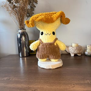 May include: A plush crochet mushroom figure with a golden-yellow cap and a pale yellow stem. The figure features black bead eyes, a stitched mouth, and is wearing brown crocheted overalls. It stands on a circular white base.