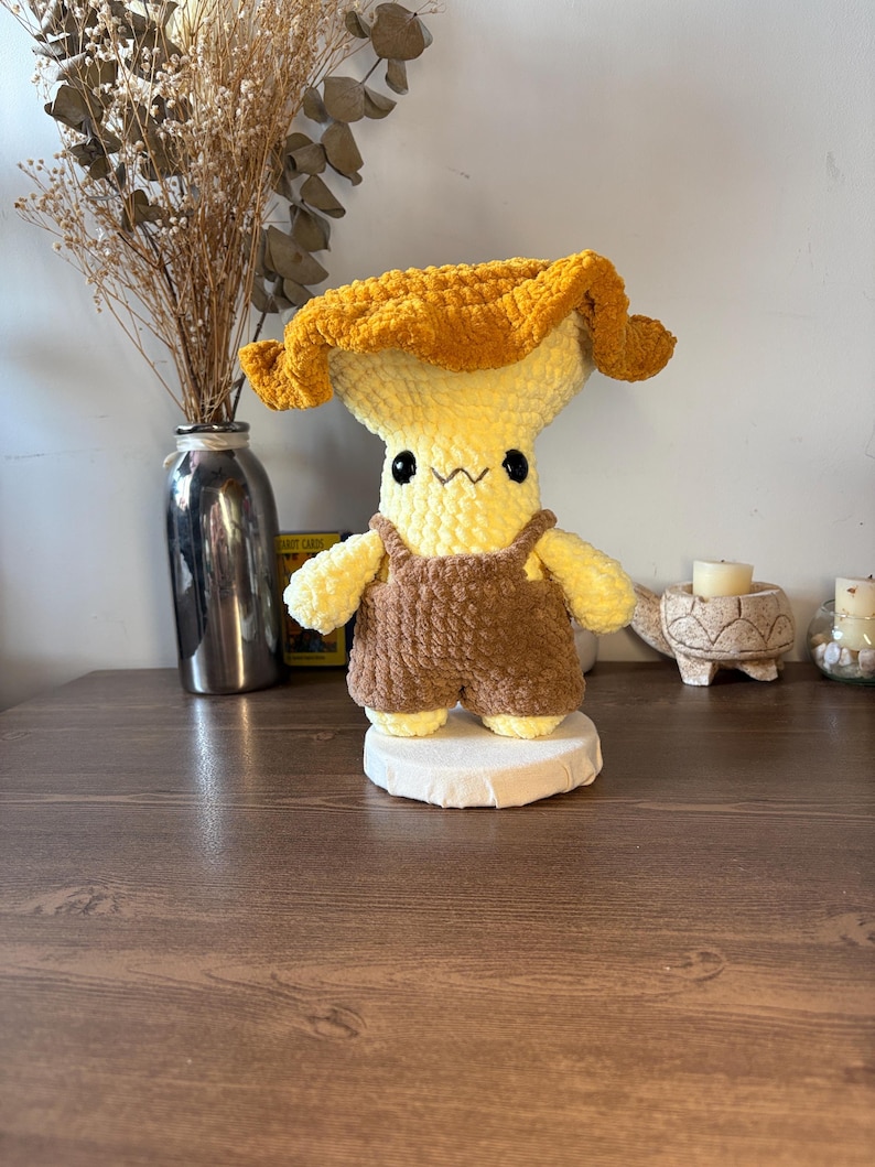 May include: A plush crochet mushroom figure with a golden-yellow cap and a pale yellow stem. The figure features black bead eyes, a stitched mouth, and is wearing brown crocheted overalls. It stands on a circular white base.