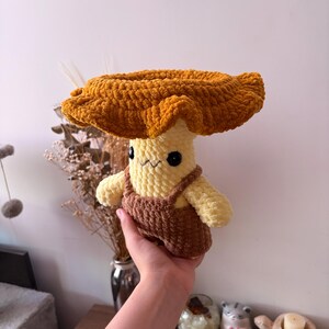 May include: A handmade crochet plush toy shaped like a mushroom with a wide, ruffled golden-brown cap and a pale yellow stem. The plush features small black safety eyes, a stitched mouth, and is wearing brown crocheted overalls with straps.