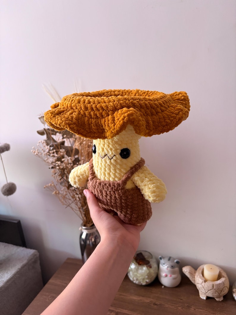 May include: A handmade crochet plush toy shaped like a mushroom with a wide, ruffled golden-brown cap and a pale yellow stem. The plush features small black safety eyes, a stitched mouth, and is wearing brown crocheted overalls with straps.