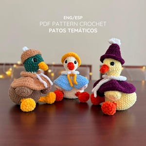以下が含まれることがあります： ENG/ESP PDF PATTERN CROCHET PATOS TEMÁTICOSという文字が上に書かれた、3体の編みぐるみのカモ。左のカモは茶色と緑色でベージュのケープを着用、中央は白で青いケープ、右は黄色で紫色のケープを着用しています。