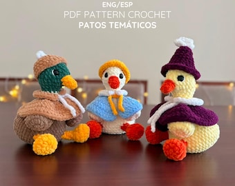 May include: Three crocheted duck figures with text above reading ENG/ESP PDF PATTERN CROCHET PATOS TEMÁTICOS. The left duck is brown and green with a tan cape, the center is white with a blue cape, and the right is yellow with a purple cape.