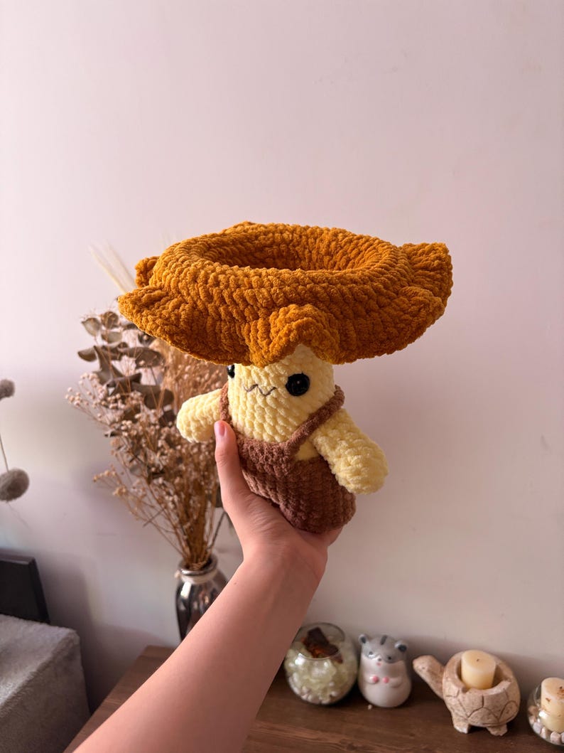 May include: A plush crochet mushroom figure with a wide, textured golden-brown cap and a pale yellow stem. The figure features small black safety eyes, a stitched mouth, and is wearing brown crocheted overalls with thin straps over its body.