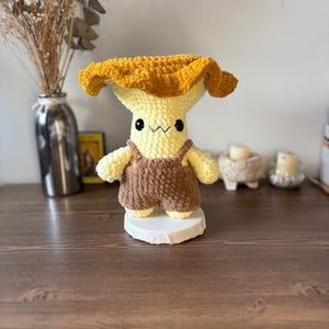 May include: A crochet plush toy shaped like a mushroom with a wide, textured golden-yellow cap and a pale yellow stem. The figure features small black bead eyes, a stitched mouth, and is wearing brown crocheted overalls with shoulder straps.