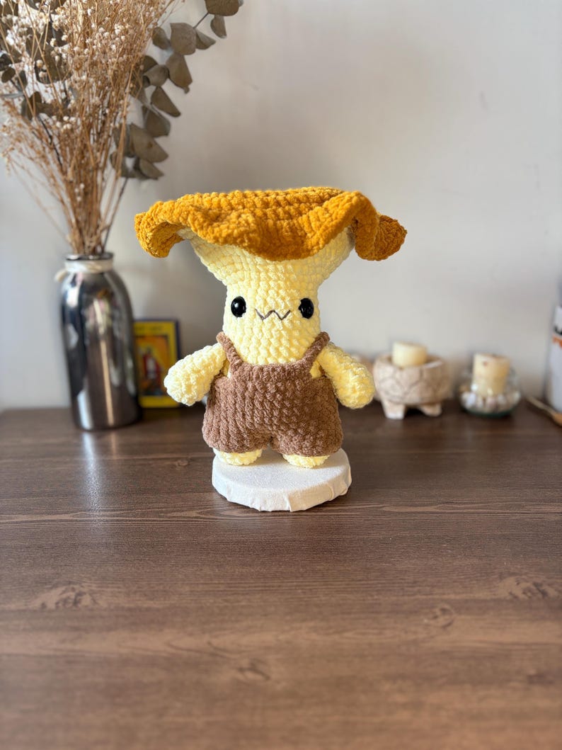 May include: A crochet plush toy shaped like a mushroom with a wide, textured golden-yellow cap and a pale yellow stem. The figure features small black bead eyes, a stitched mouth, and is wearing brown crocheted overalls with shoulder straps.