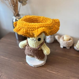 May include: A crocheted plush figure featuring a cream-colored body with black embroidered eyes and a small mouth, wearing brown yarn overalls. The figure is topped with a large, wide-brimmed, textured mustard yellow hat made of soft yarn.