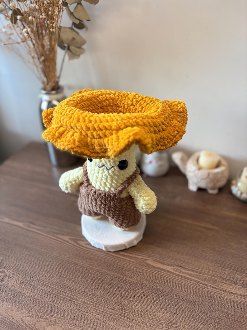 May include: A crocheted plush figure featuring a cream-colored body with black embroidered eyes and a small mouth, wearing brown yarn overalls. The figure is topped with a large, wide-brimmed, textured mustard yellow hat made of soft yarn.