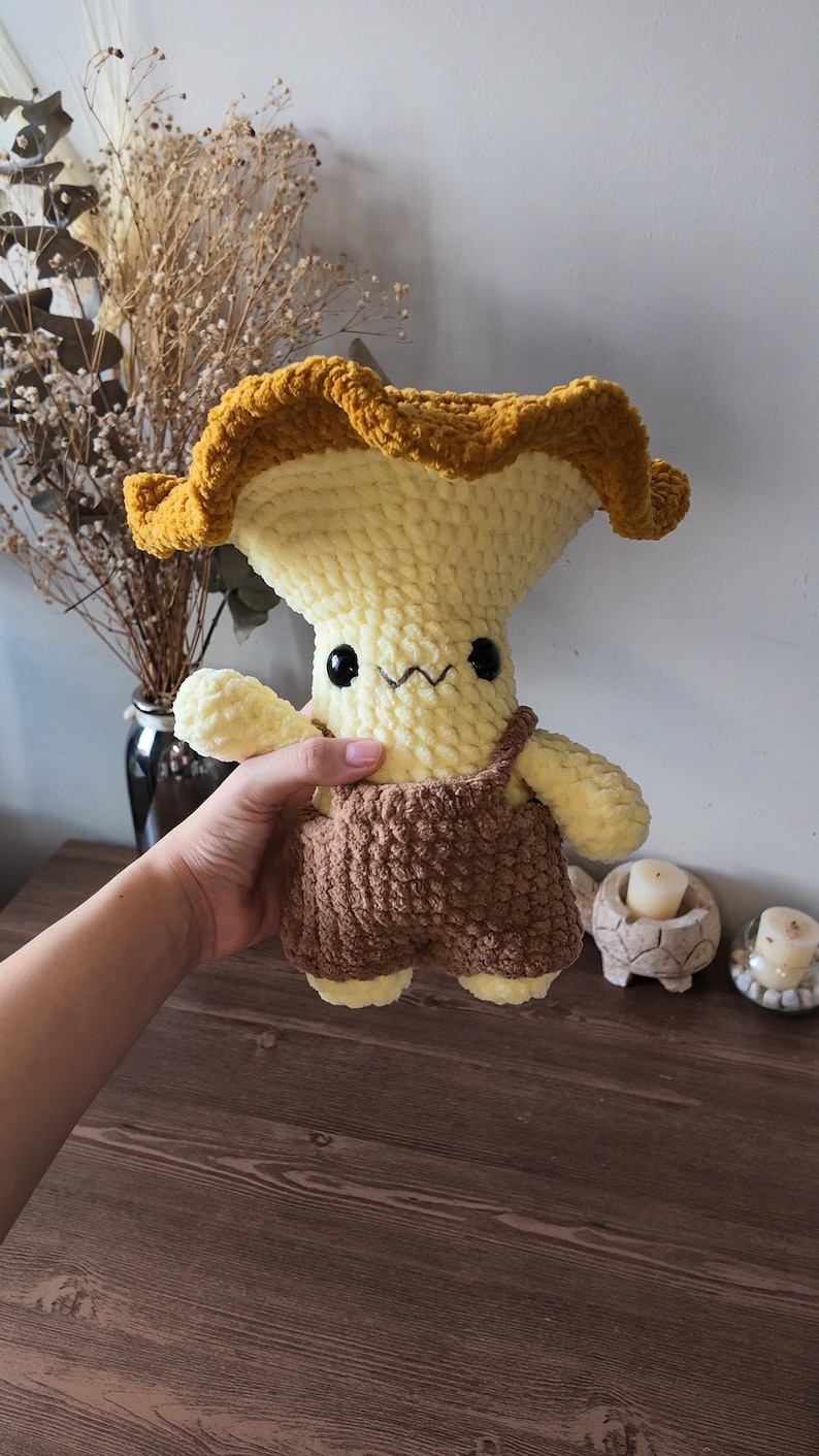 May include: A plush crochet mushroom character with a wide, ruffled golden-brown cap and a pale yellow stem. The figure features small black bead eyes, a stitched mouth, and is wearing brown textured overalls with visible shoulder straps.