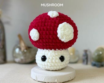 May include: A handmade crochet mushroom with a red cap and white spots, atop a white stem. The mushroom has black eyes and is displayed on a small, round, white platform. Text on the image reads "PDF PATTERN CROCHET MUSHROOM".