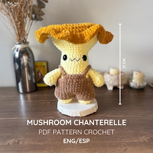 May include: A crochet mushroom doll with a golden-yellow cap and a pale yellow stem, wearing brown overalls. The doll has black bead eyes and a stitched mouth. A vertical line indicates a height of 21 cm. Text reads: MUSHROOM CHANTERELLE PDF PATTERN CROCHET ENG/ESP.